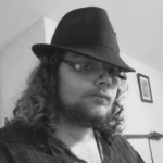 Black and white headshot for Seth Lamb. White male with long curly hair, beard and mustache, wearing glasses and a fedora.