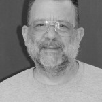 Black and white headshot for Cal Jones. White male with short gray hair, beard and mustache, and wearing glasses.