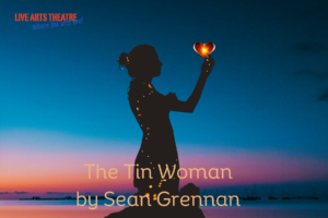 Show logo for The Tin Woman. A landscape rectangle with a blue and pink sunrise in the background. There is a black silhouette of a woman holding a glowing heart in the foreground. In the upper right corner is the Live Arts logo. The logo says Live Arts Theatre in red stencil font and Where the Arts live in blue cracked font below it at an angle sloping upward from left to right. At the bottom center, it says The Tin Woman by Sean Grennan in gold font.