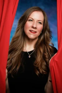 Color headshot. A caucasian woman with shoulder length hair who is wearing a black shirt and standing between red curtains. There is a blue background behind her.