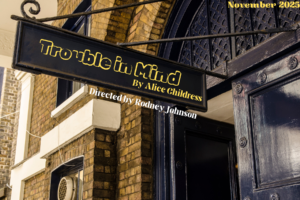 Show logo for Trouble in Mind. A brick building with a black sign. On the sign, the words Trouble in Mind by Alice Childress are printed in yellow font. Below the sign, the words Directed by Rodney L. Johnson are printed in white font. In the top right corner, the words November 2025 are printed in yellow font.